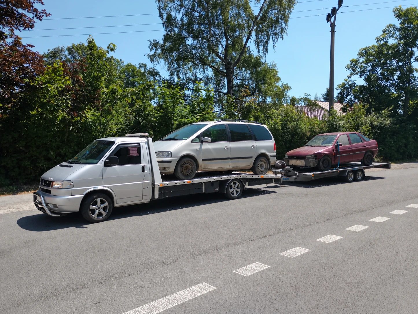 Strieborná odťahová VW T4 s VW Sharan na plošine a Fordom Escort na prívese