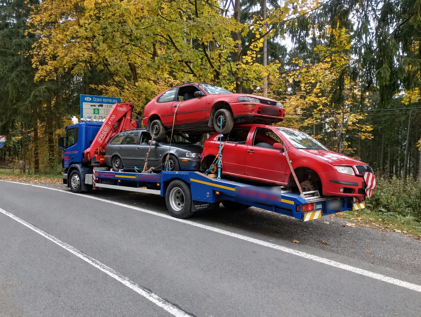 Odťahový nákladiak s hydraulickou rukou veziaci tri staré autá naraz, v pozadí jesenný les